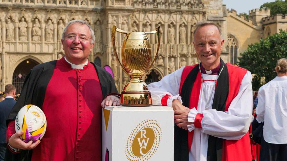 Two people wearing red and black religious robes stand beside a gold trophy. The man on the left is holding a rugby ball. There is a cathedral in the background.