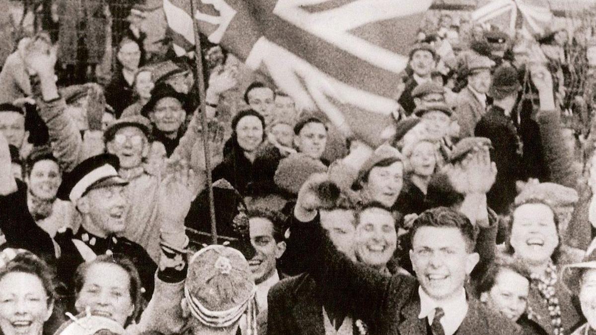 Black and white picture of people smiling as they celebrate the liberation of Guernsey on 9 May 1945. A large Union Flag is waving above the crowd.