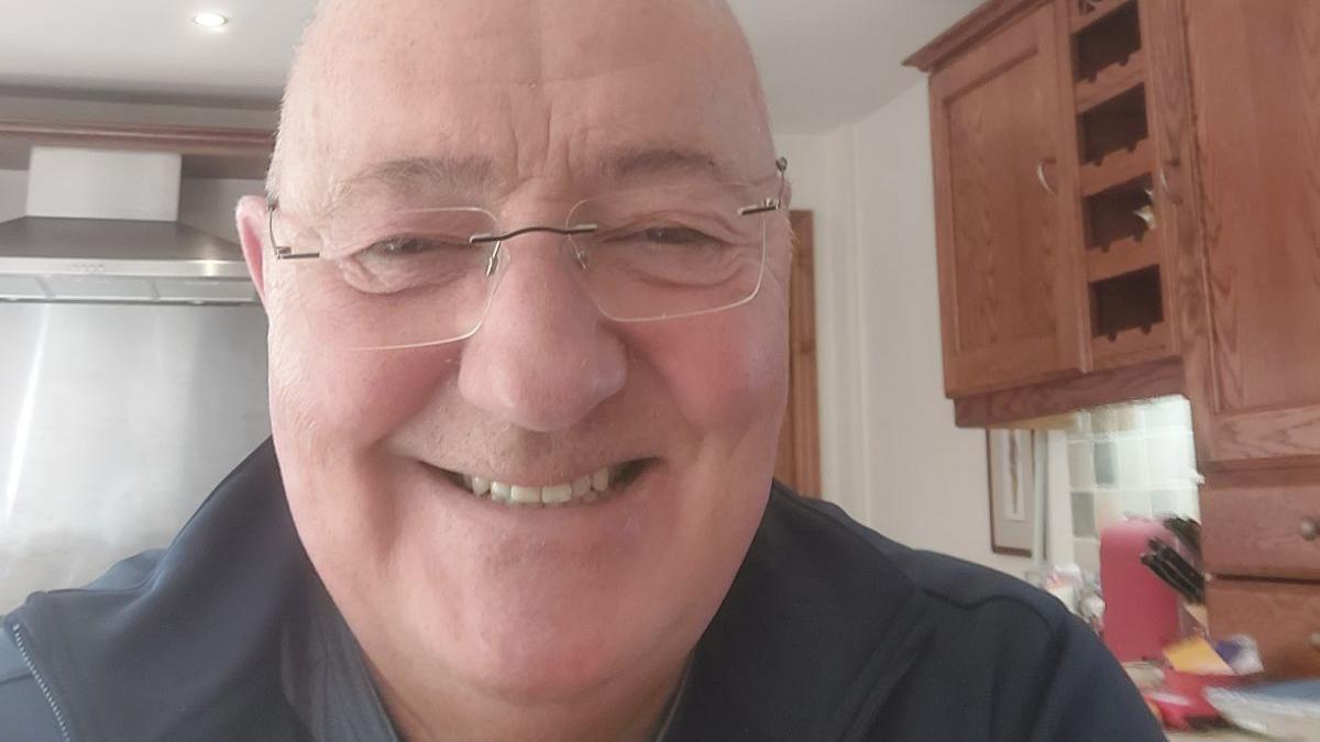 Robin Davies, bald-headed and wearing glasses and a black shirt, smiles and faces the camera in his kitchen