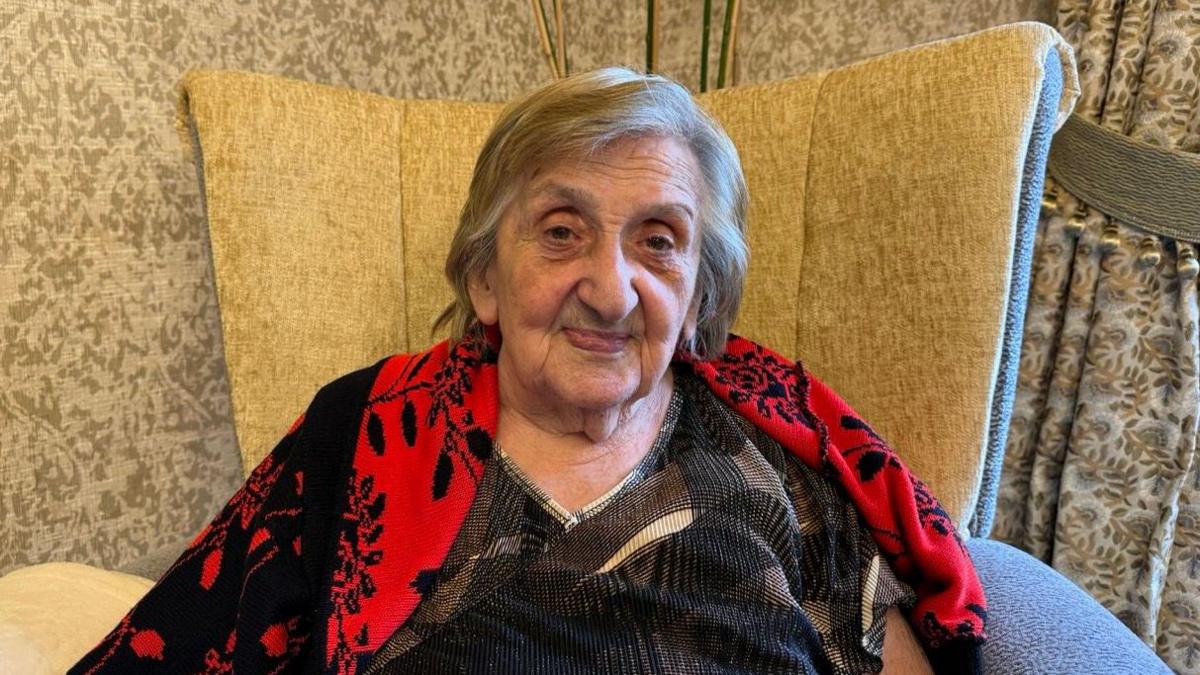 A woman sat in a big beige chair. She is smiling and has a black and red scarf draped over her shoulders. 