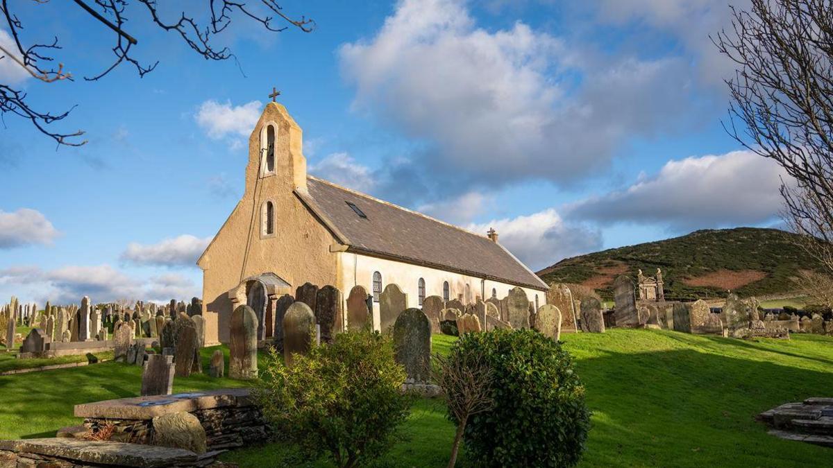 A hill-top view of Kirk Maughold church, dedicated to the saint, surrounded by gravestones on green grass banks, with a hill in the background