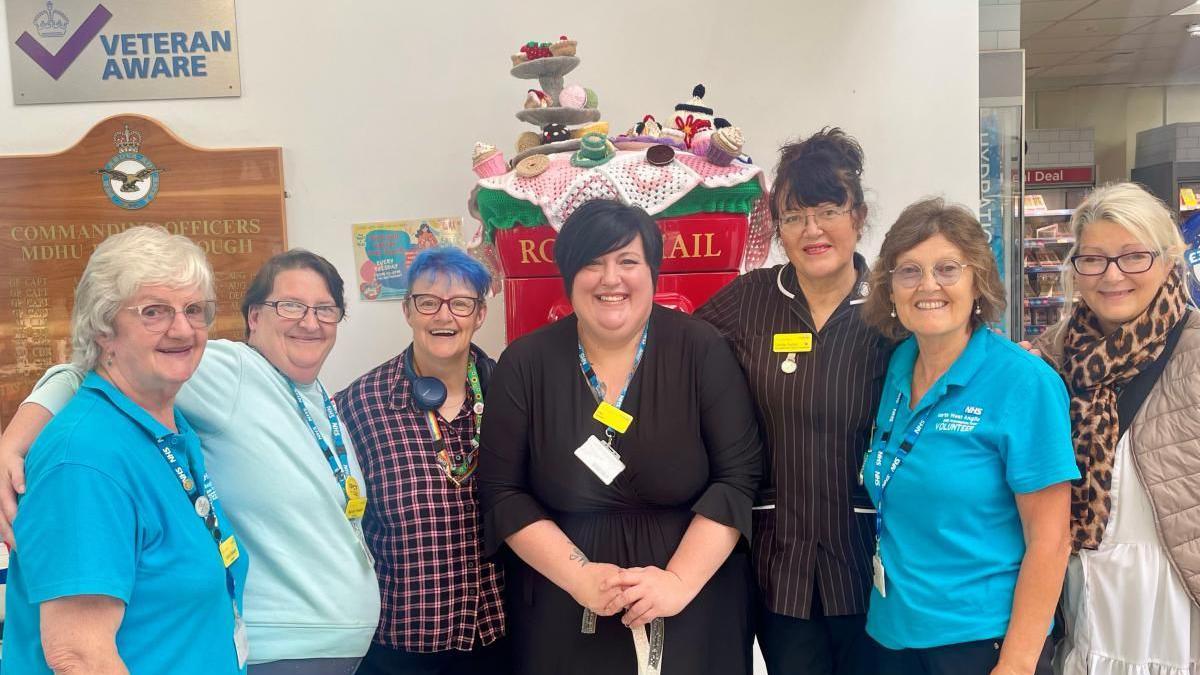 Members of the knitting group- seven women- some wearing blue T-shirts- smiling for camera in front of the post box topper