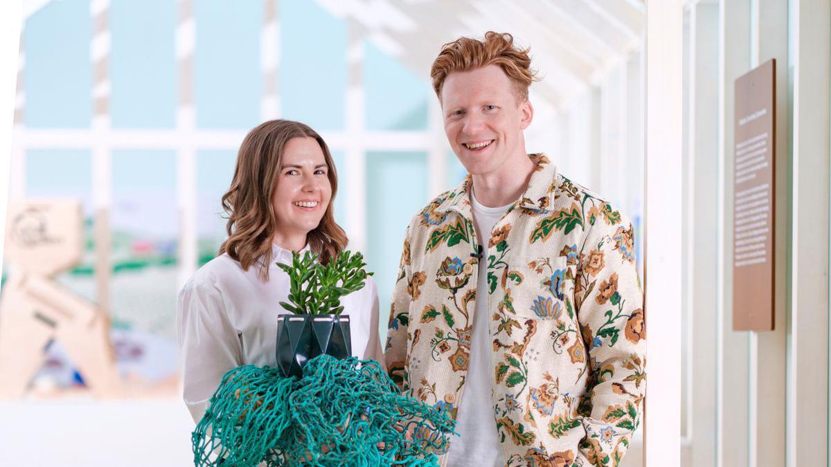 A woman and a man smile standing side by side in a bright conservatory. The woman holds a POTR pot and a bright blue fishing net. The woman had shoulder-length brown hair and the man had short ginger hair and wears a floral patterned shirt and a white t-shirt underneath