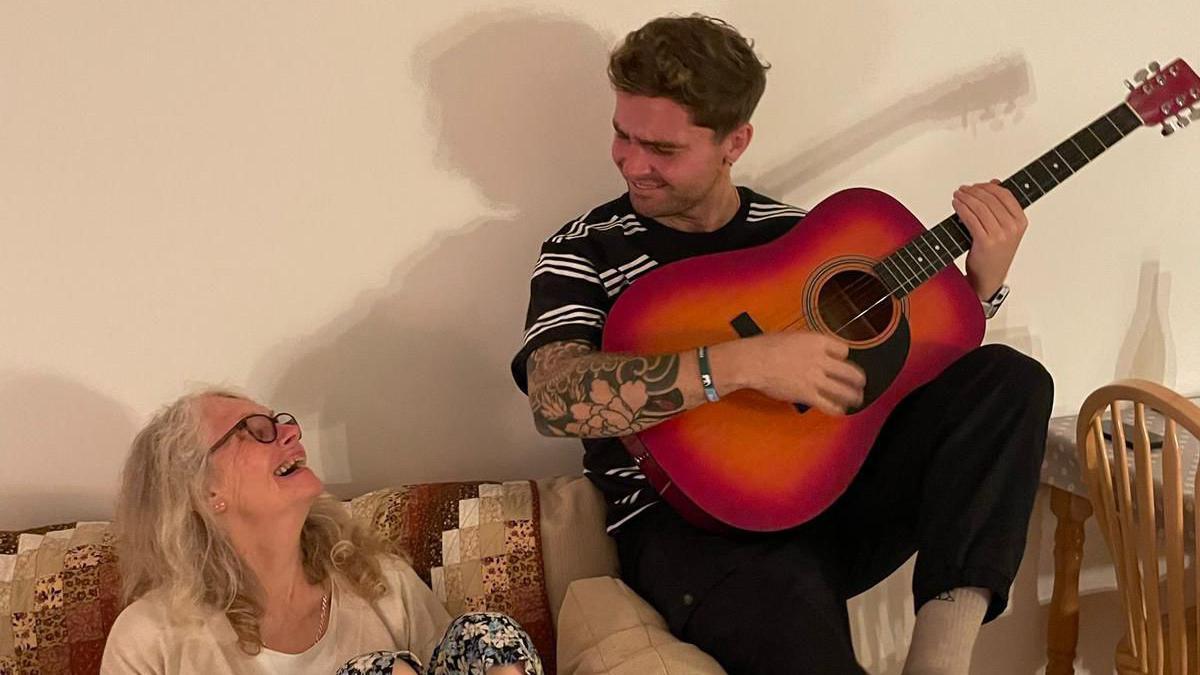 Sam King sitting on the edge of a sofa while holding an orange guitar. He is looking down at his mum who is sitting on the sofa and looking back at him. They are both smiling. 