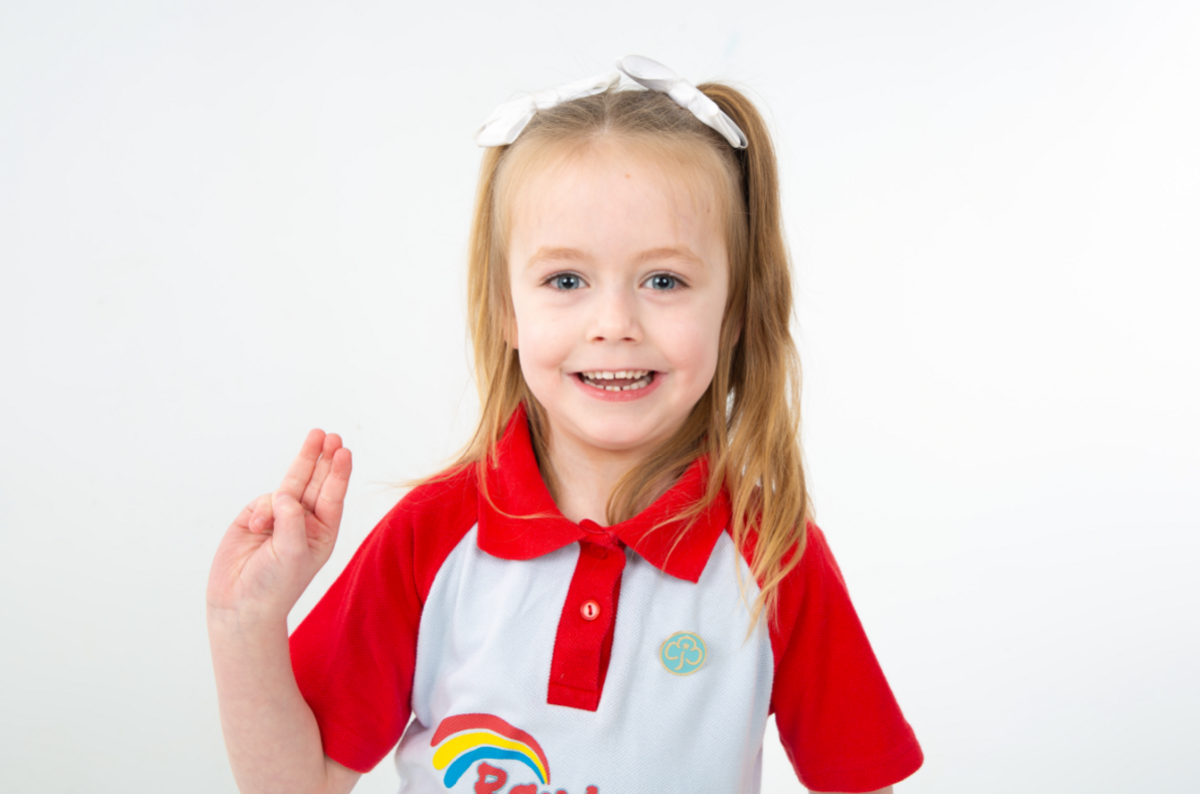Merch fach mewn crys polo coch a gwyn. Mae ganddi rhubanau gwyn yn ei gwallt melyn. 