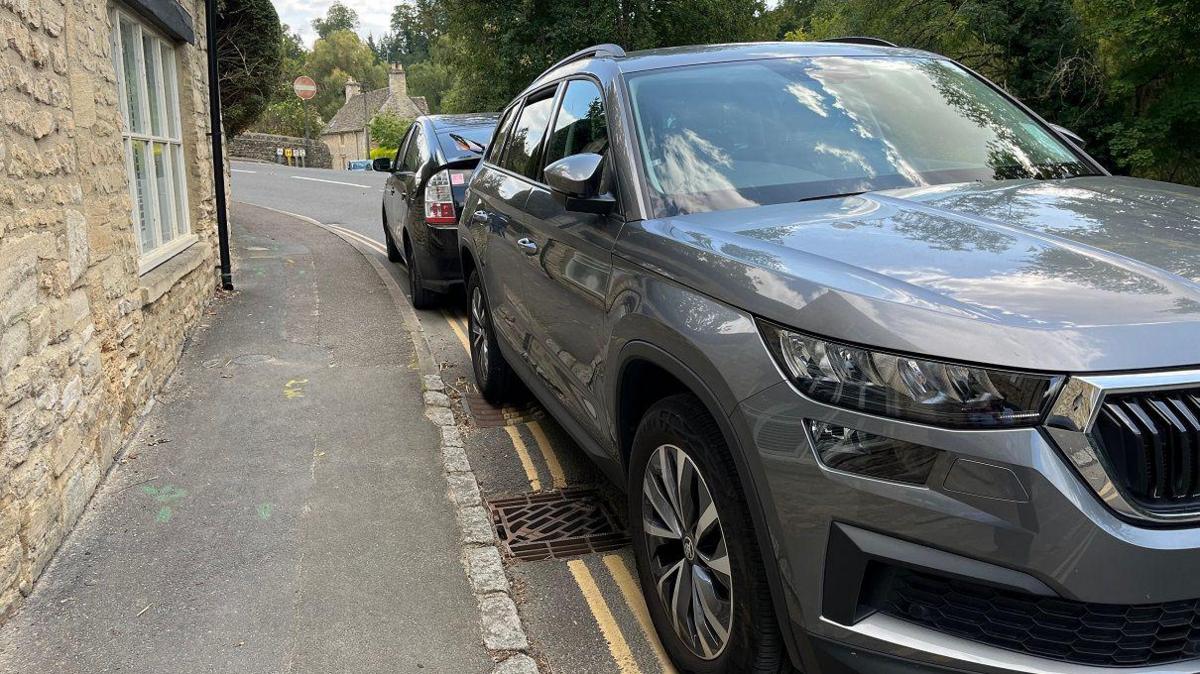 Two cars parked on double yellow lines on a road in the Cotswolds village of Bibury. One is a large grey Skoda and there is a smaller black car parked behind it.