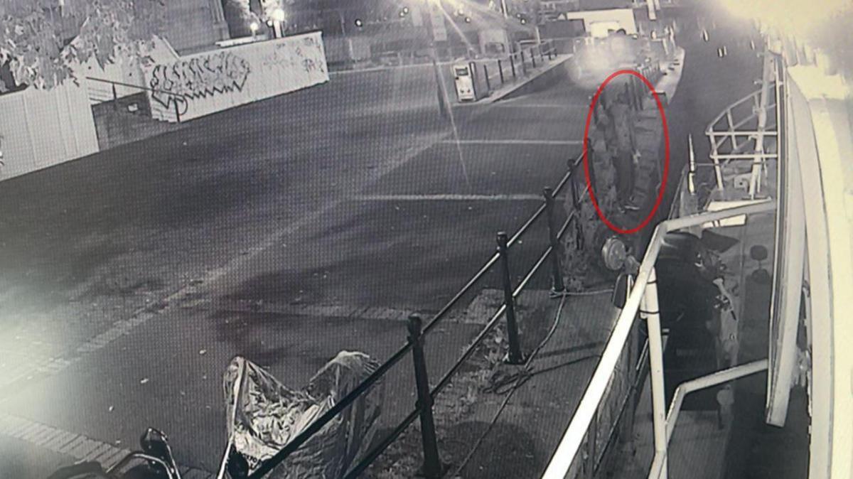 Black and white CCTV footage of  person standing past a set of black railings, right by the Lady Kate. The area its lit up by street lights.