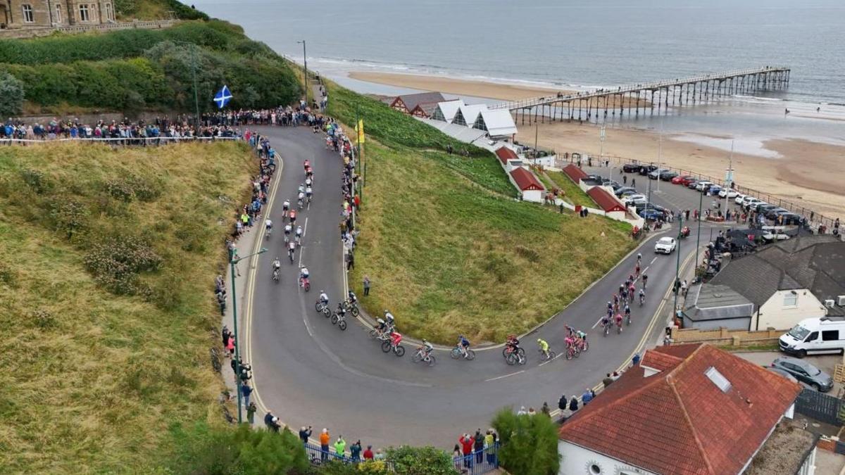 A high-up view of cyclists taking part in the Tour of Britain negotiating a steep road winding up from a beach. The roadside is lined with people. At the top right hand side a pier extends from the beachfront into the sea, which is at low tide