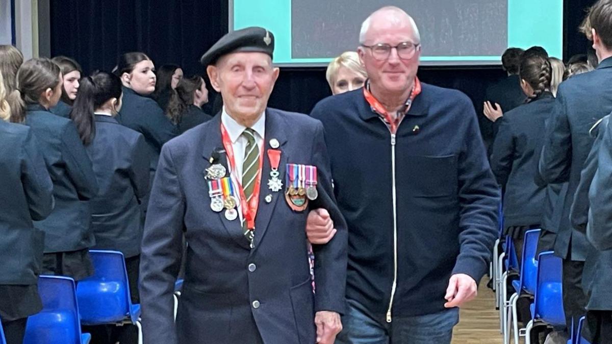 Two men walking down the aisle of a school hallway. One is wearing a dark blazer with multiple military medals and a beret. The other man is dressed in a zip-up jacket and jeans, walking alongside and supporting the other man by holding his arm. In the background, a large projection screen displays a message and an image. Either side of the men are male and female students dressed in  dark coloured school uniform. 