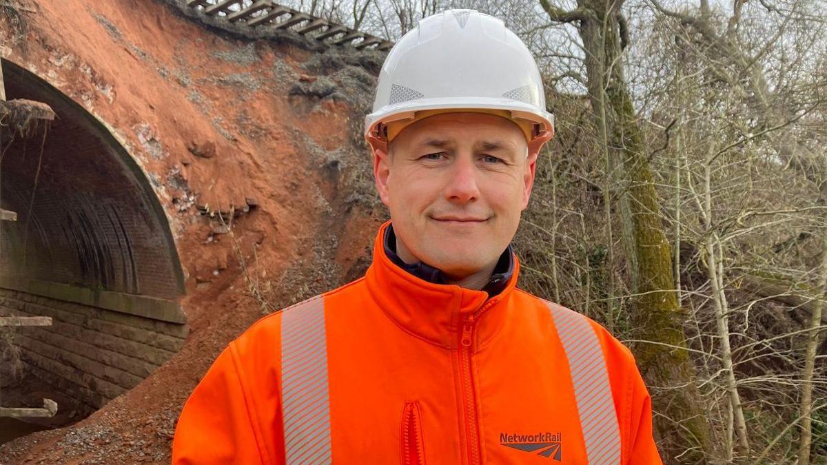 A man is wearing a white hard hat and a bright orange Network Rail coat. He is standing in front of a railway bridge that has land missing from the top of it, with tracks suspended over the edge