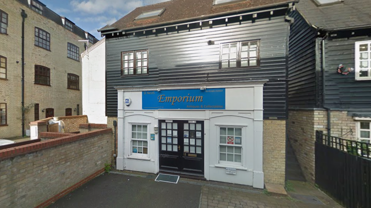 The entrance to a shop named 'Emporium', featuring a black door with glass panes and a small accessibility ramp. The building has a white brick lower facade and a black wooden upper facade, with windows on both levels. 
