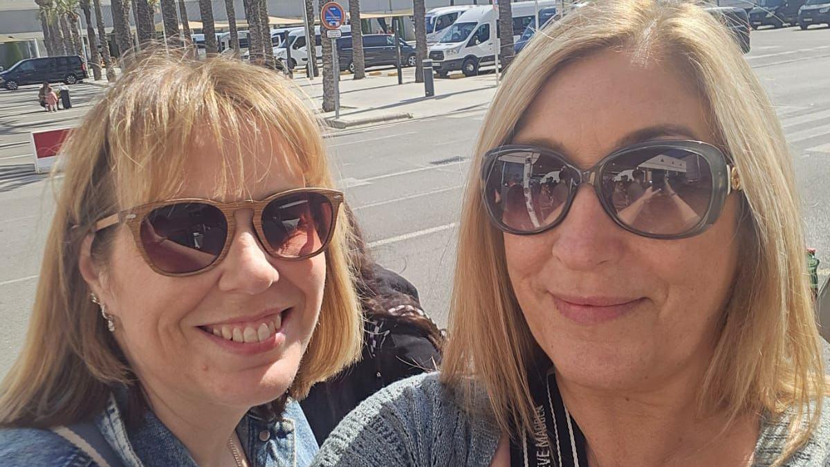 Two blonde women - both wearing sunglasses - smiling on a sunny day. A carpark and palm trees are in the background.