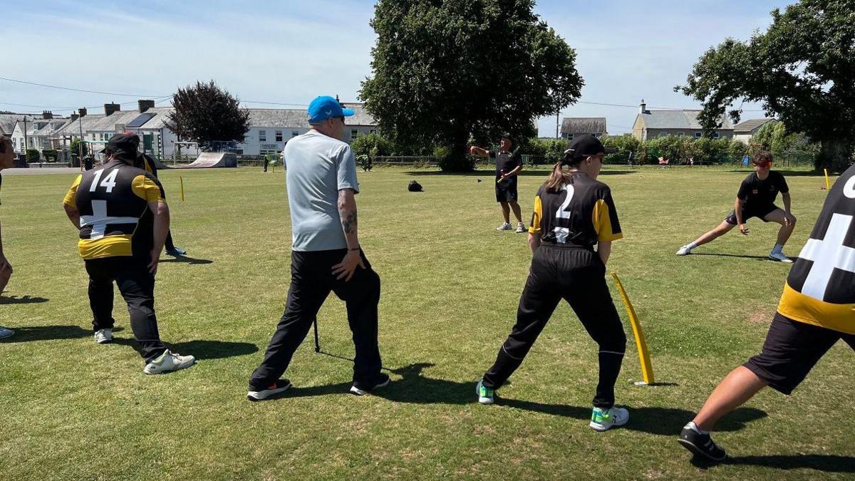 Five men and one woman are on a cricket pitch doing warm-up exercises and stretches. Five of them are wearing a sports strip which are black, gold and yellow with St Piran flags and a team number on them.