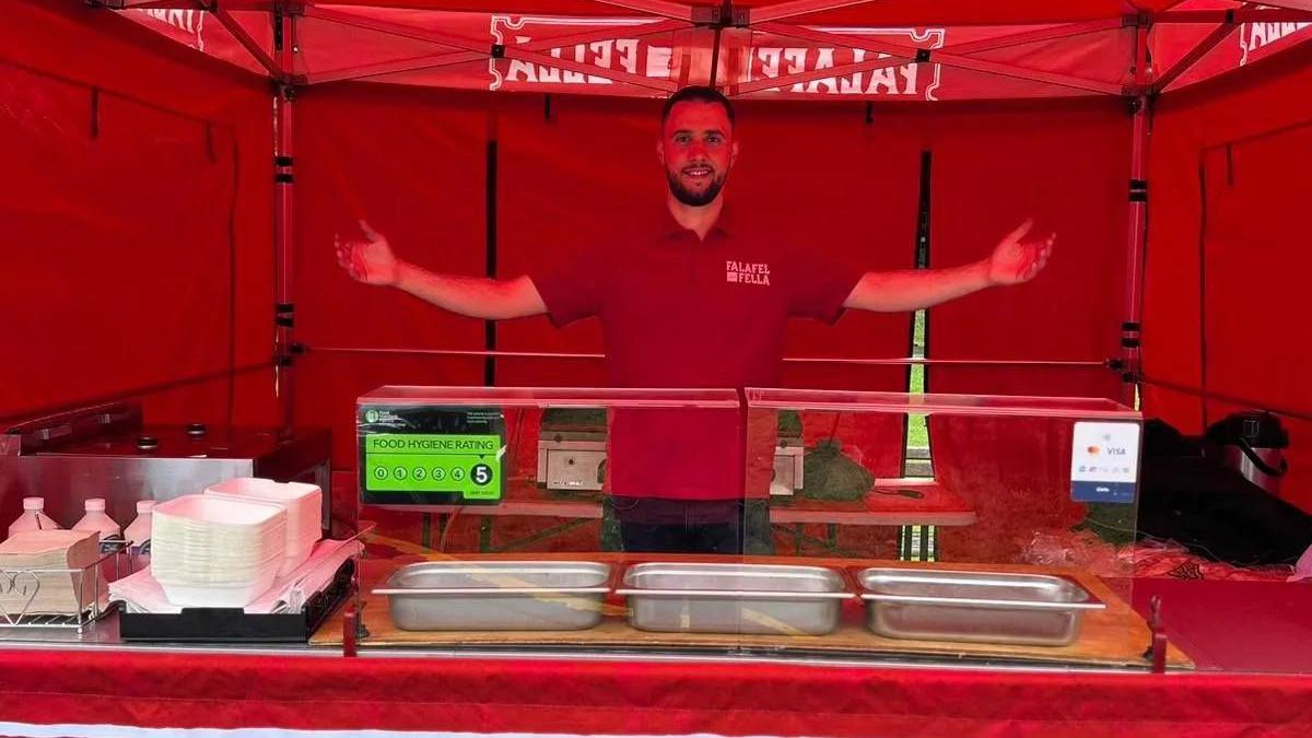 A man which short black hair and a short black beard stands looking happy behind a street food stall. He has his arms outstretched and wears a red T-shirt.