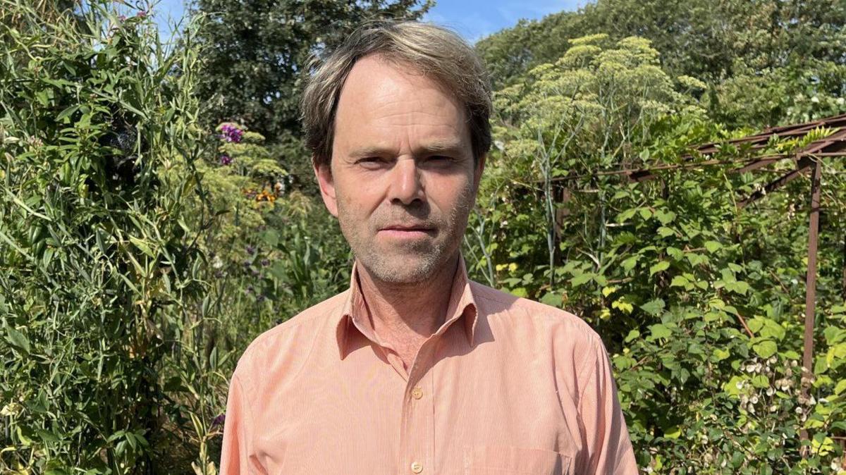 Environmental campaigner Professor Rupert Read in his Norfolk garden. He is looking directly at the camera and there is a lot of green foliage behind him. 