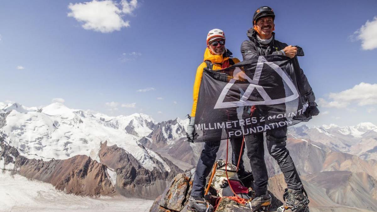 Mr Jackson and his friend standing on the peak of a mountain, holding out a black flag which says "Millimetres to Mountains Foundation". They are on the highest mountain visible, with the others far below and covered in snow. The men are wearing black trousers, hiking boots, a yellow coat, and helmets. They both look happy and proud of their accomplishment.