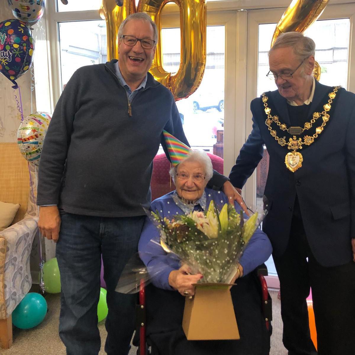 Two men and a woman at a birthday party, with balloons in the background. The man on the left has grey hair and glasses, and he is wearing a blue jumper and jeans. The man on the right has grey hair and glasses, and he is wearing large gold ceremonial necklaces over a dark jacket. The woman, who is sitting down, has grey hair and glasses. She is wearing a stripey cone-shaped party hat and a purple jumper.