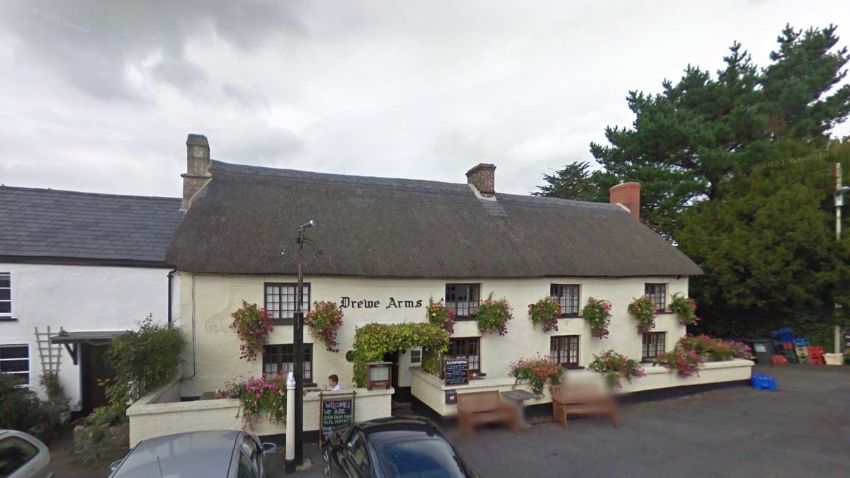 Exterior of The Drewe Arms. The cream pub has a thatched roof. It has eight windows across the front of the building. A red sign on the building states Drewe Arms. A number of benches are in front of the pub. 