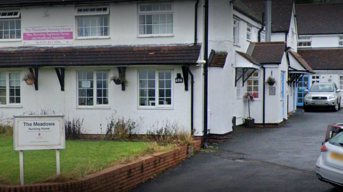 The outside of a white rendered building with brown roof tiles. A car park is visible to the right hand side. On the left is a small patch of lawn with a sign that says The Meadows Nursing Home