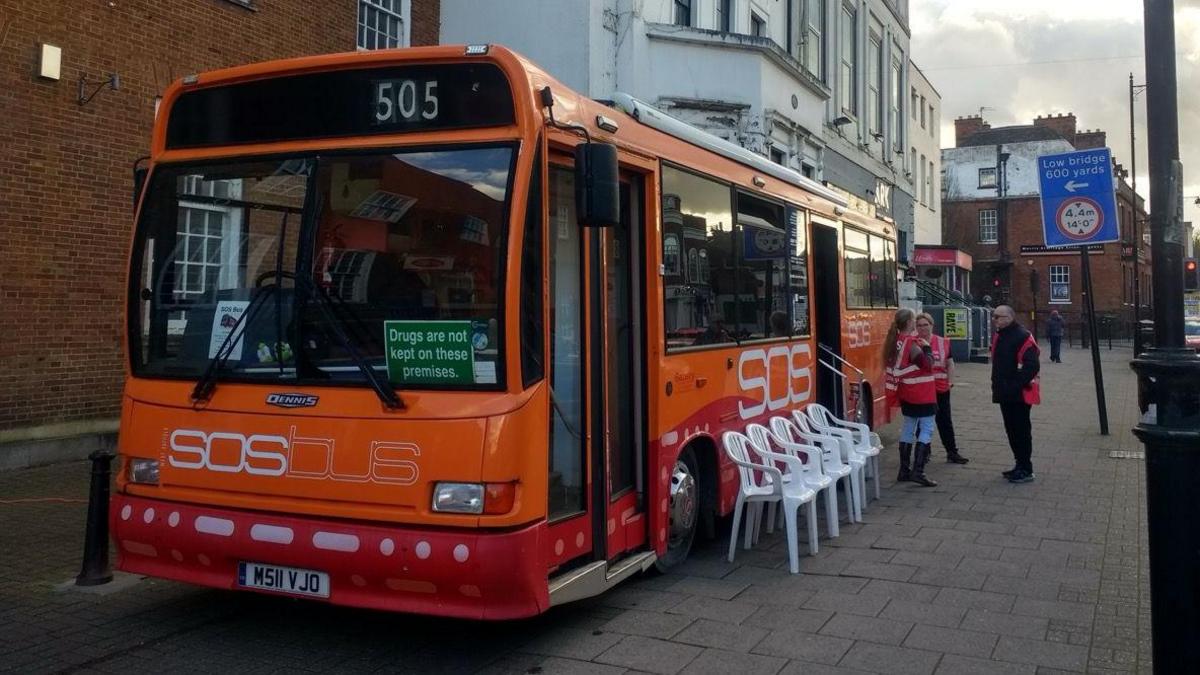 A single-decker orange bus is parked on pavement in a town centre. It is orange and red and has the words SOS Bus written on its front. Volunteers in red bibs stand outside and several white plastic chairs have been placed along the side of the bus.