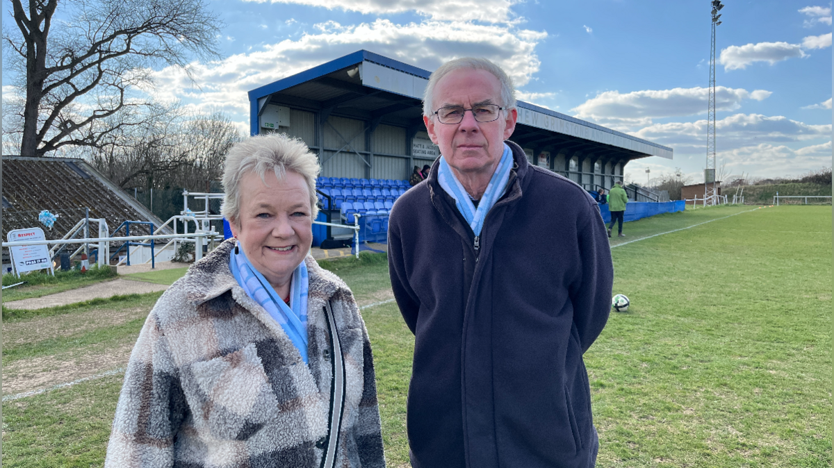A man and woman stand on a football pitch on a sunny day. The man, Phil Grimstone, has grey hair and glasses. He is wearing a dark-coloured fleece, is not smiling and is holding his arms behind his back. The woman, Caroline Schilt, is smiling and is wearing a thick, checked coat. She has grey short hair.