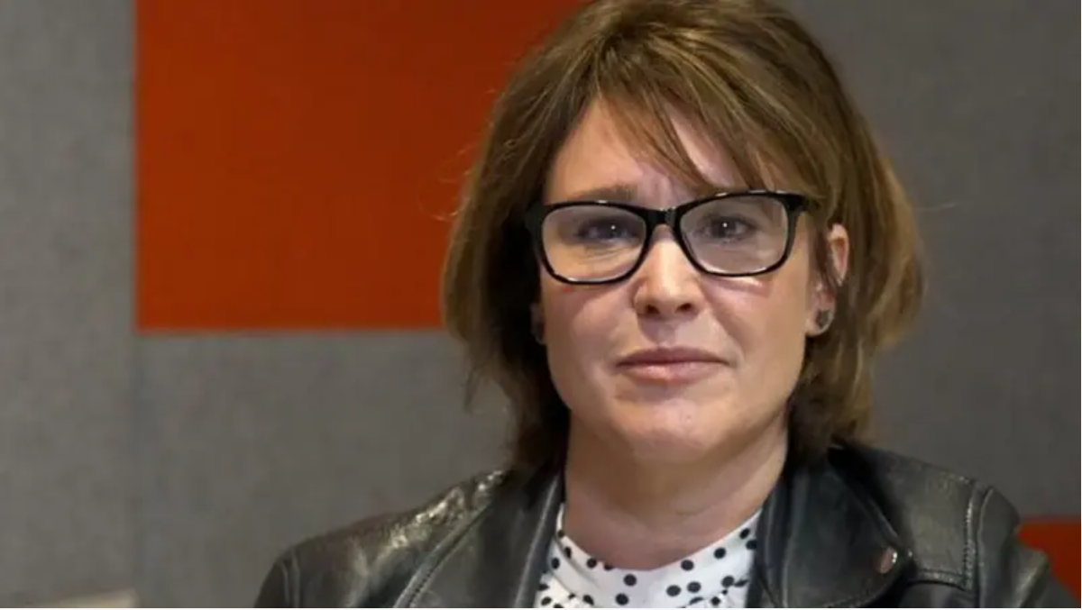 Carly has light brown shoulder length hair and is wearing black glasses, a leather jacket and is looking at the camera whilst sitting in an office