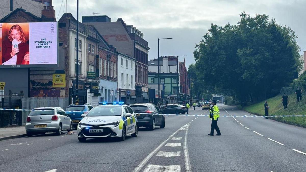 General view of the southbound carriageway of Bristol Street in Birmingham. A police cordon is stretching across the street with a police officer in the foreground and four officers dressed in black standing on a sloped grass verge. A police car is also visible in the foreground