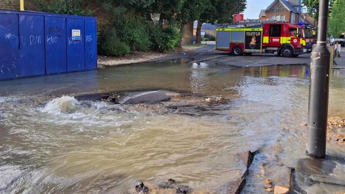 A large amount of water floods a suburban street. A fire engine is visible at one end of the road. Water is escaping through a sink hole in the road.