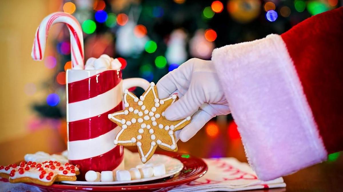 The outstretched arm of a person dressed as Father Christmas holding a star-shaped cookie. There is a red and white striped candle in the background and other treats.