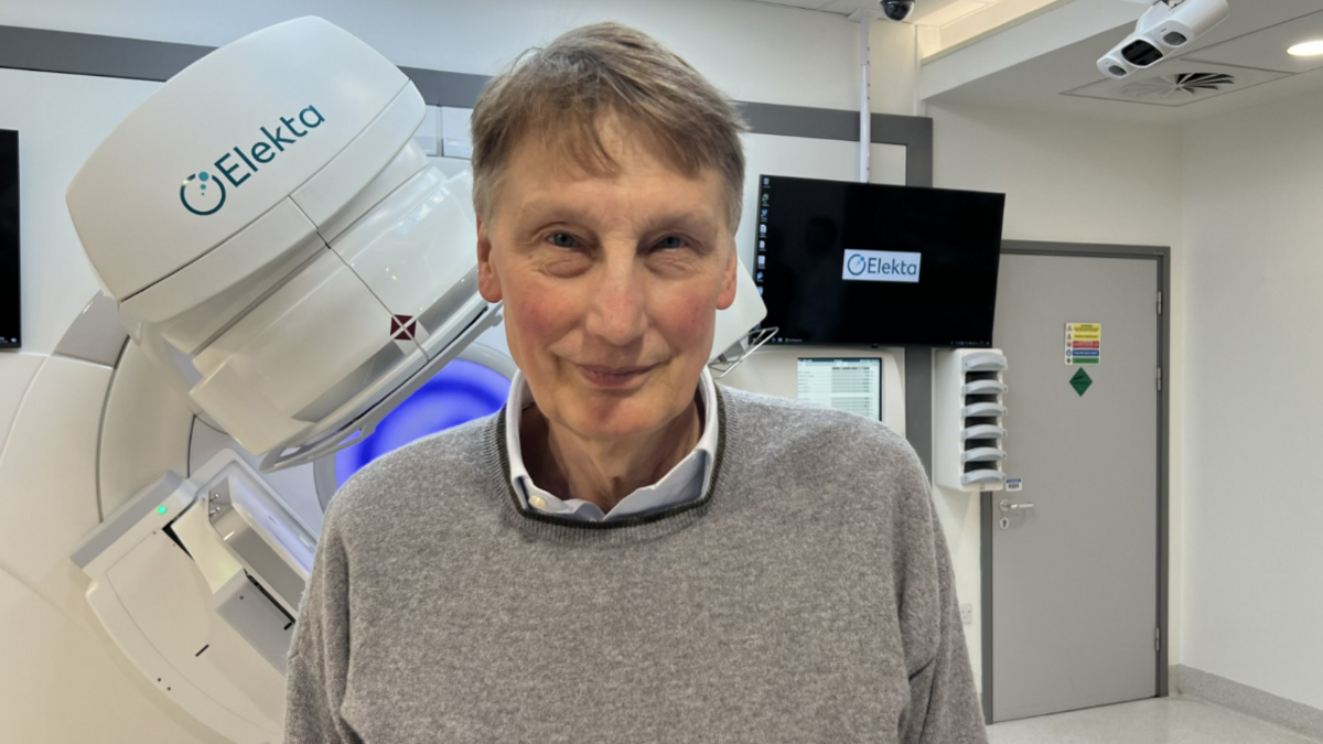 Prof Adrian Crellin wearing a grey jumper and blue shirt stood in front of cancer treatment equipment at a hospital 