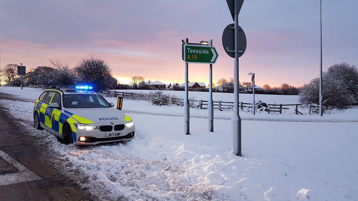 A police car with its blue lights on is parked on the side of a road in a field which is full of snow. A roadside nearby says 'Teesside A19'.