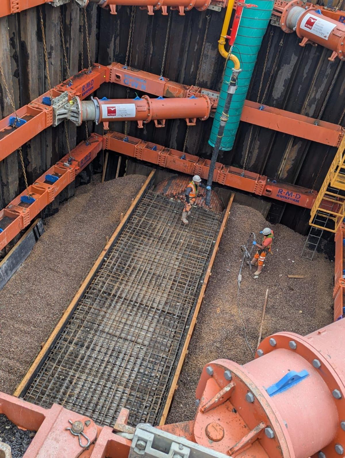 Orange pipes at the bottom of the sinkhole. There are two men in orange suits working at the bottom. There is a grate at the bottom levelling out the floor. 
