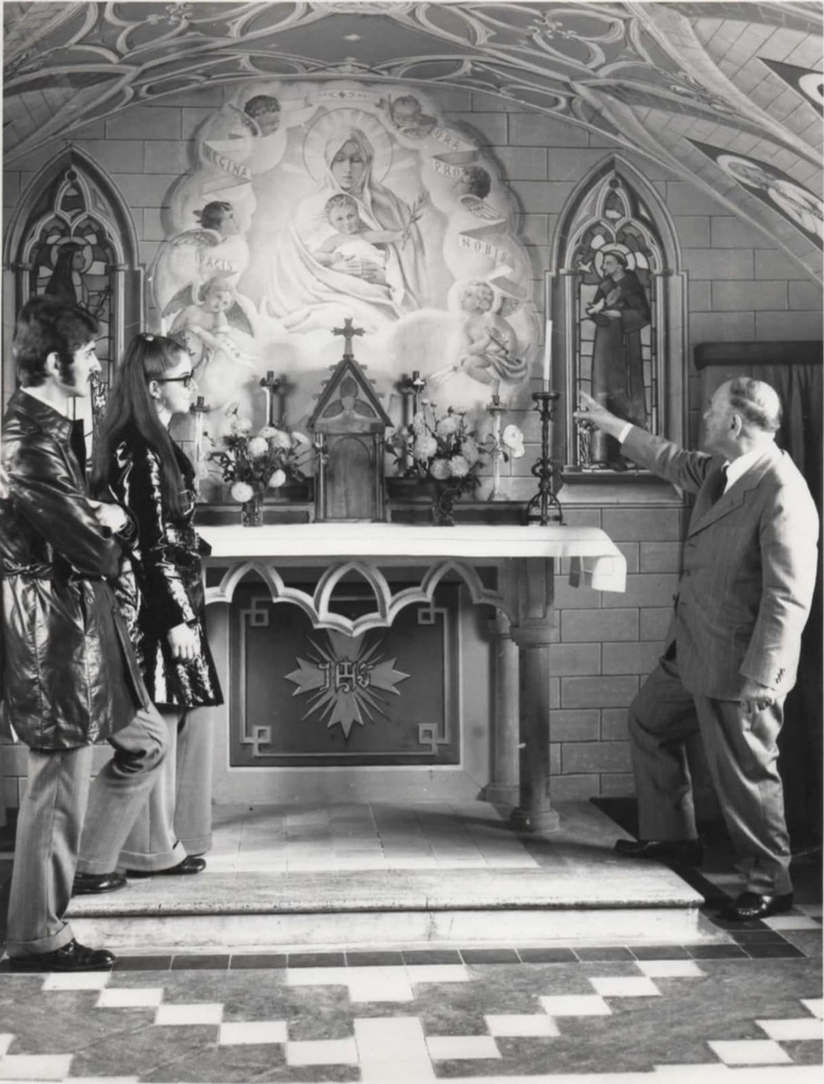 Two young adults in jeans and dark coats stand on one side of an alter, talking to an older man in a suit. He is pointing to the painting of Christ and the Virgin Mary.