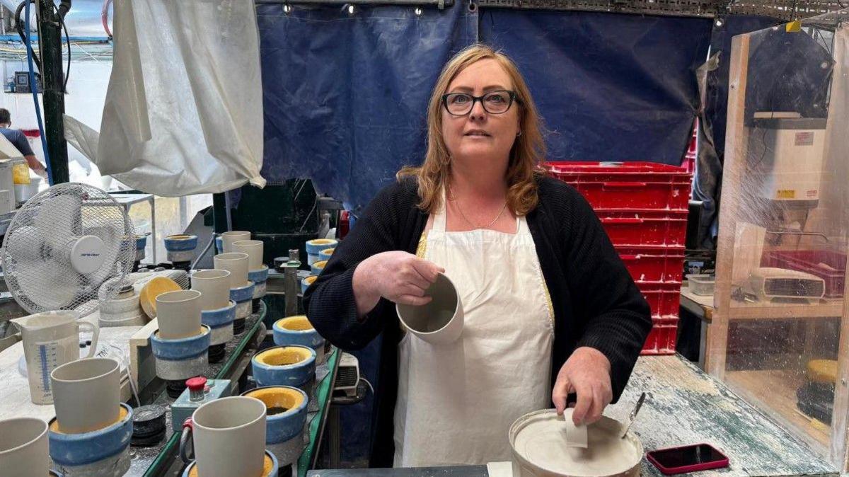 A woman with shoulder-length brown hair is wearing glasses. She has a white apron on and is holding a cup. At least a dozen mugs are on the left of the photo.