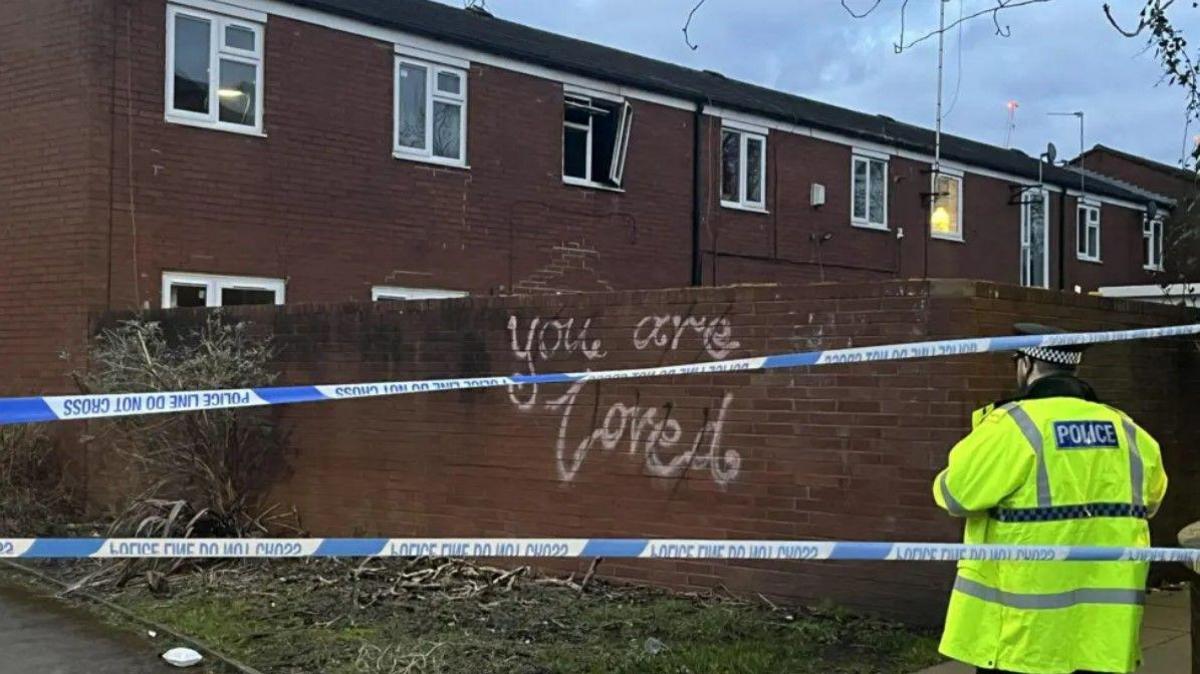 A blackened, burnt-out window can be seen on the first floor of the terraced block. Behind blue-and-white police tape and an officer wearing a yellow hi-vis jacket, graffiti reading "You are Loved" has been sprayed in white paint on a wall at the rear of the block.

