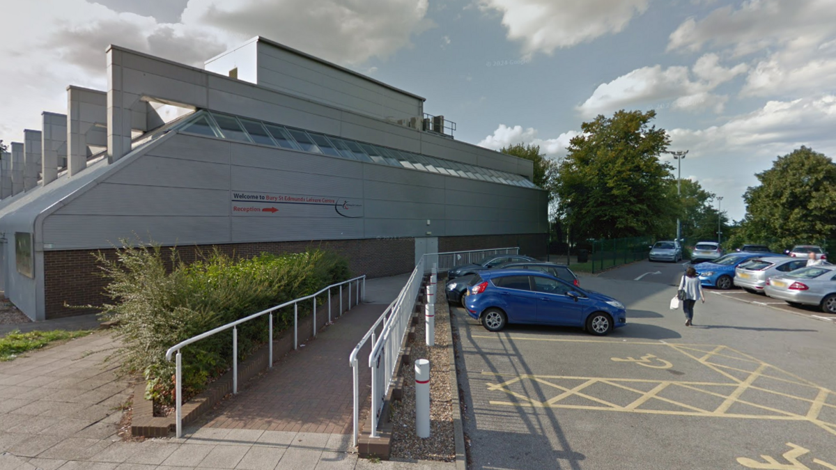 The outside of Bury St Edmunds leisure centre. A large white building with red and black writing on, Welcome to Bury St Edmunds Leisure Centre, with a sign which says reception and an arrow pointing to the right side of the building. There is also a car park outside the leisure centre with disabled parking bays and a ramp up to the leisure centre with white railings.