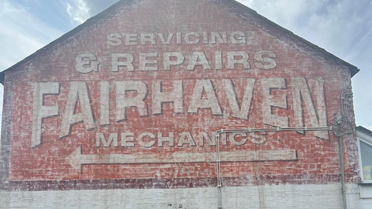 Side of a building with a faded painted advertisement reading "Servicing & Repairs FAIRHAVEN MECHANICS" and an arrow pointing left.