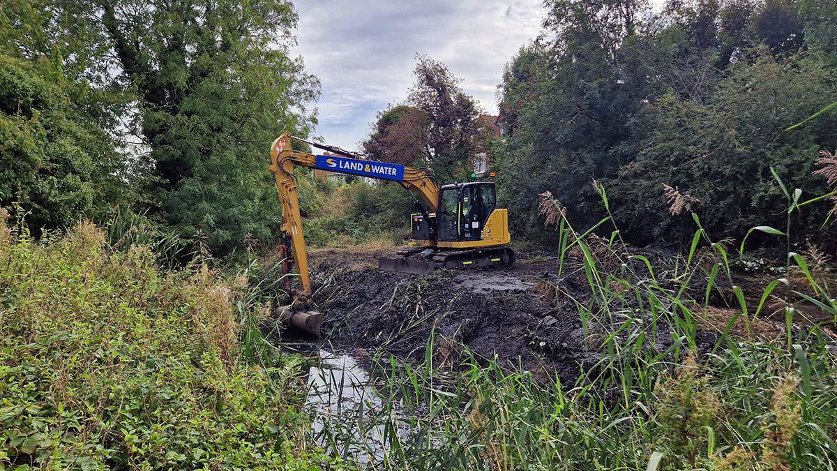 A yellow digger amongst the greenery of a nature reserve digging out silt from a waterway