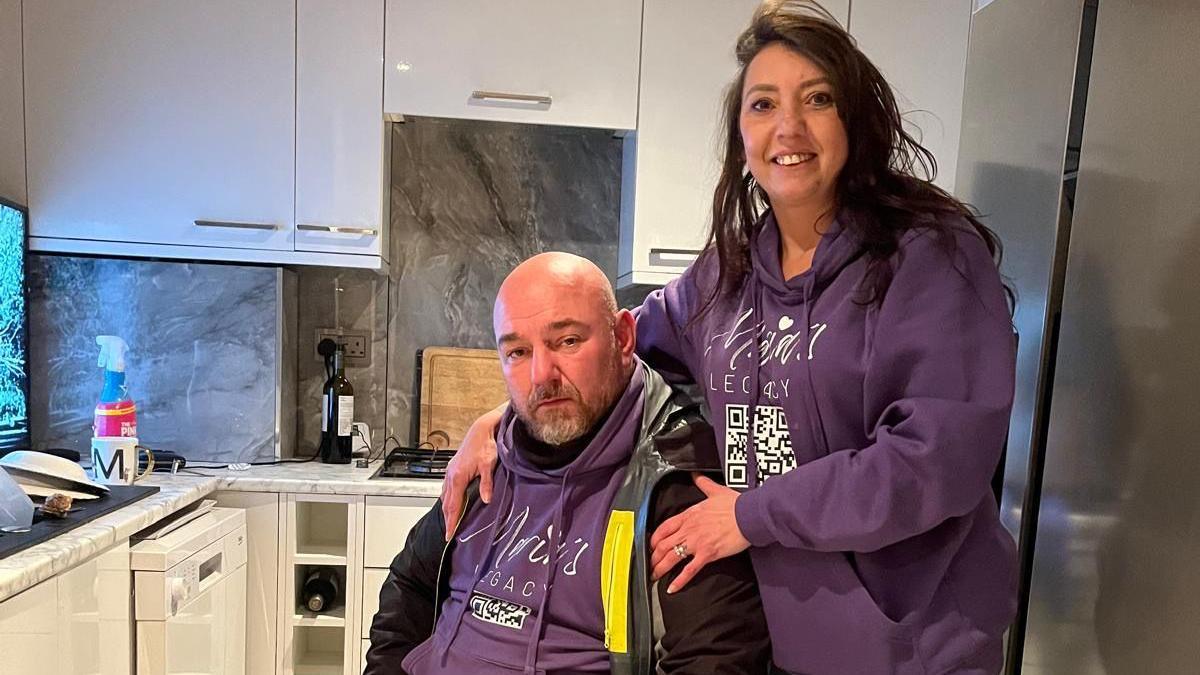 Santino and Michelle pictured in purple jumpers with the words "Maria's legacy" in white writing with a QR code printed at the bottom of the jumper. Santino is sat down and also wearing a black jacket while Michelle is stood up and behind him, holding onto his shoulders and smiling. They are pictured in a grey kitchen with white cupboards 