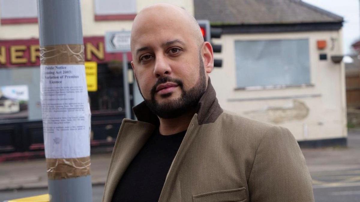 Nathan is wearing a brown jacket with the collar turned up and a black top underneath. He is stood by a lamppost on a street with a building behind him. He has a beard and a shaved head.