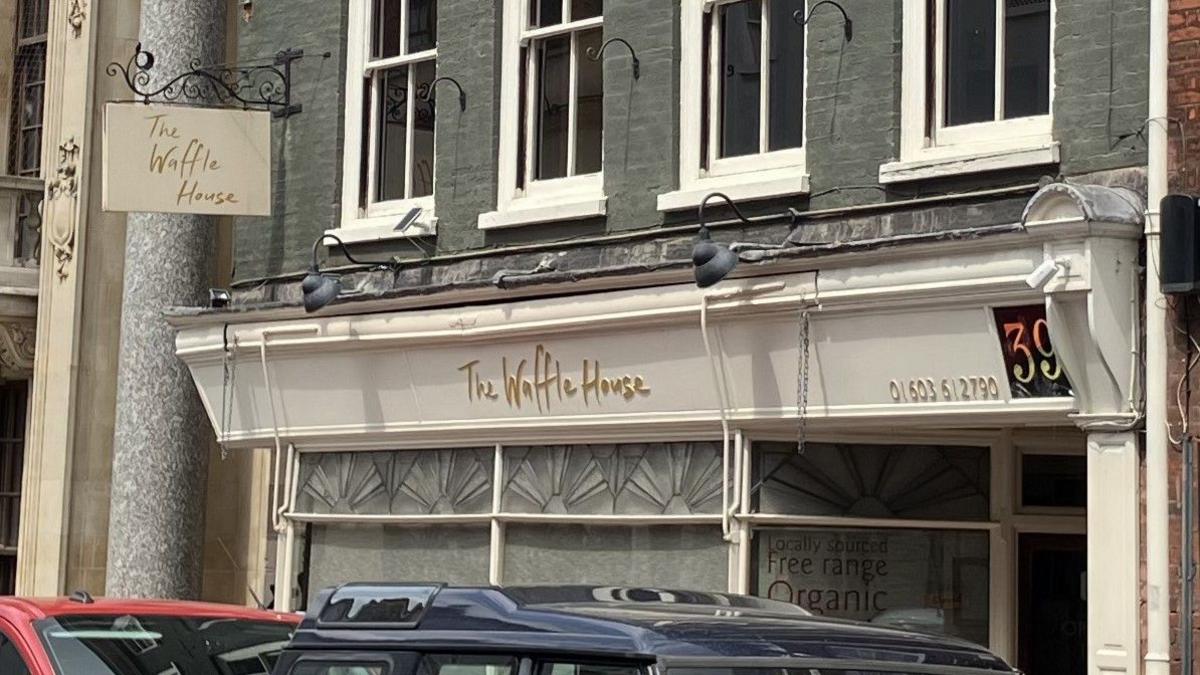 The outside of a restaurant called The Waffle House shows a sign hanging outside and its curtains drawn due to it being closed