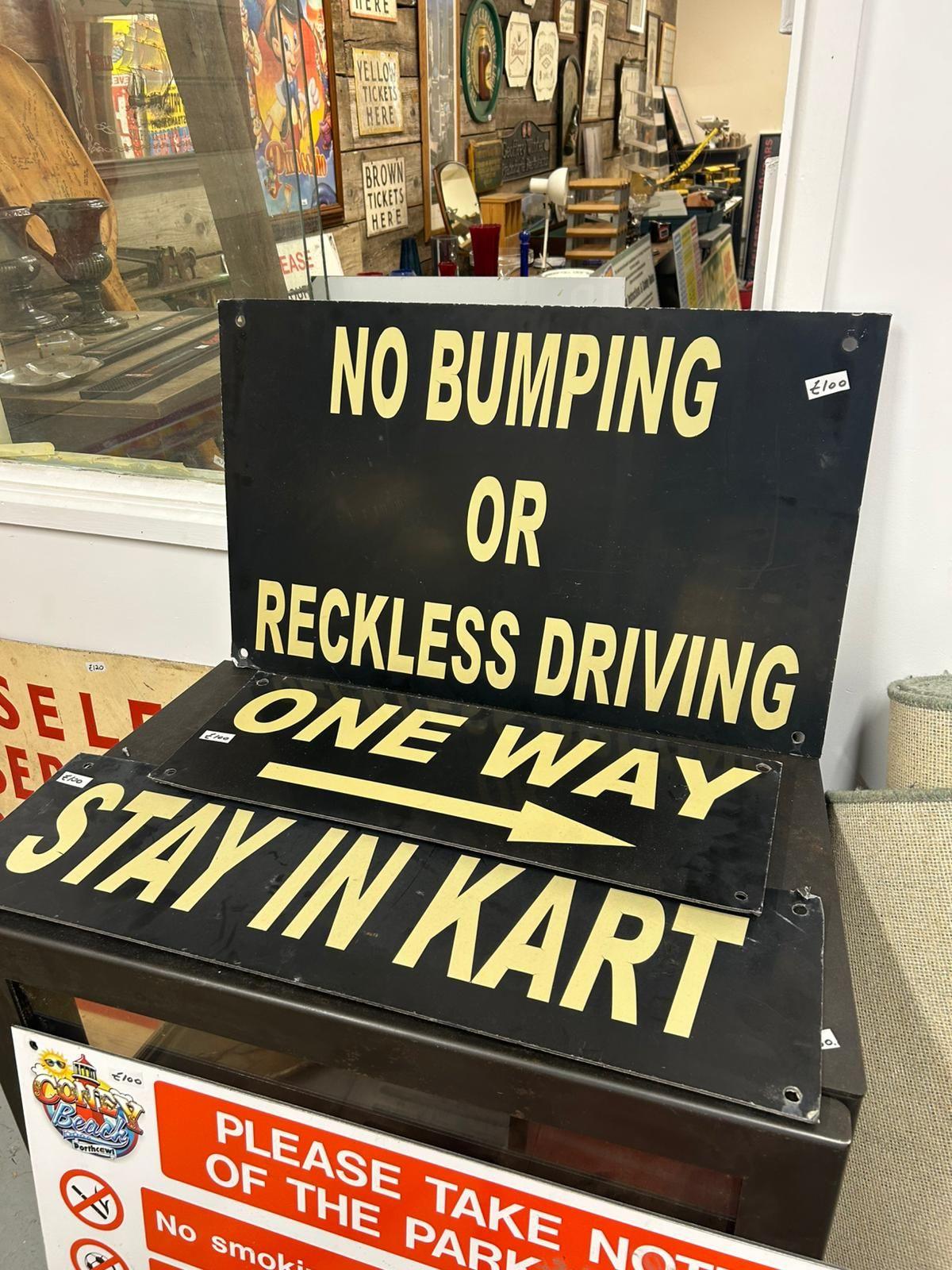 Multiple black signs with pale yellow writing, they read (top to bottom): 'no bumping or reckless driving' and 'one way' with an arrow pointing to the right beneath it' and 'stay in kart'