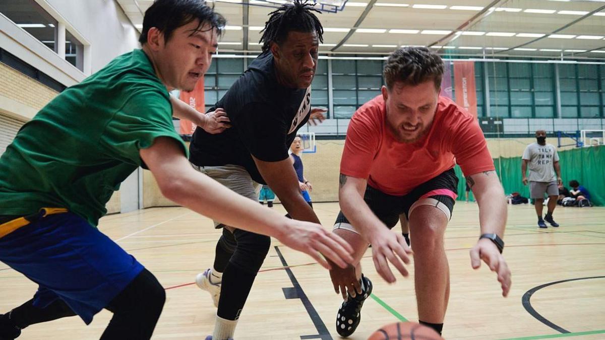 Three men in sports clothing in a wood-floored sports hall, all reaching for an orange ball on the ground