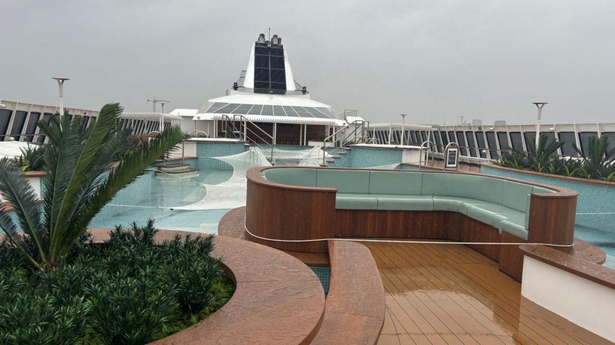 A wooden seating area with blue cushions is on the deck. Behind this is a pool with steps into it.