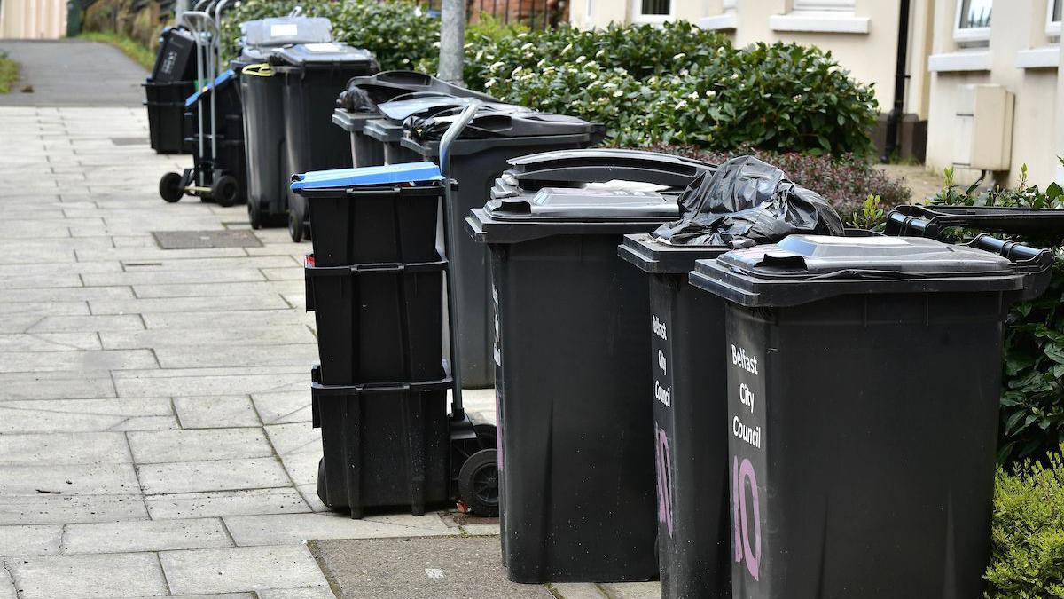 A row of black bins. Some of them are open with black bin bags overflowing. There is a stack of smaller black bin boxes. On the front of the bins it says Belfast City Council in white writing.