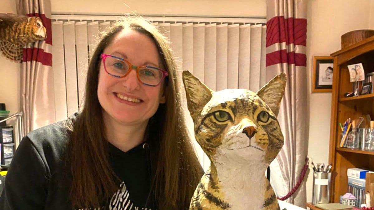 A woman with glasses smiling while standing next to a sculpture of a cat made using newspaper