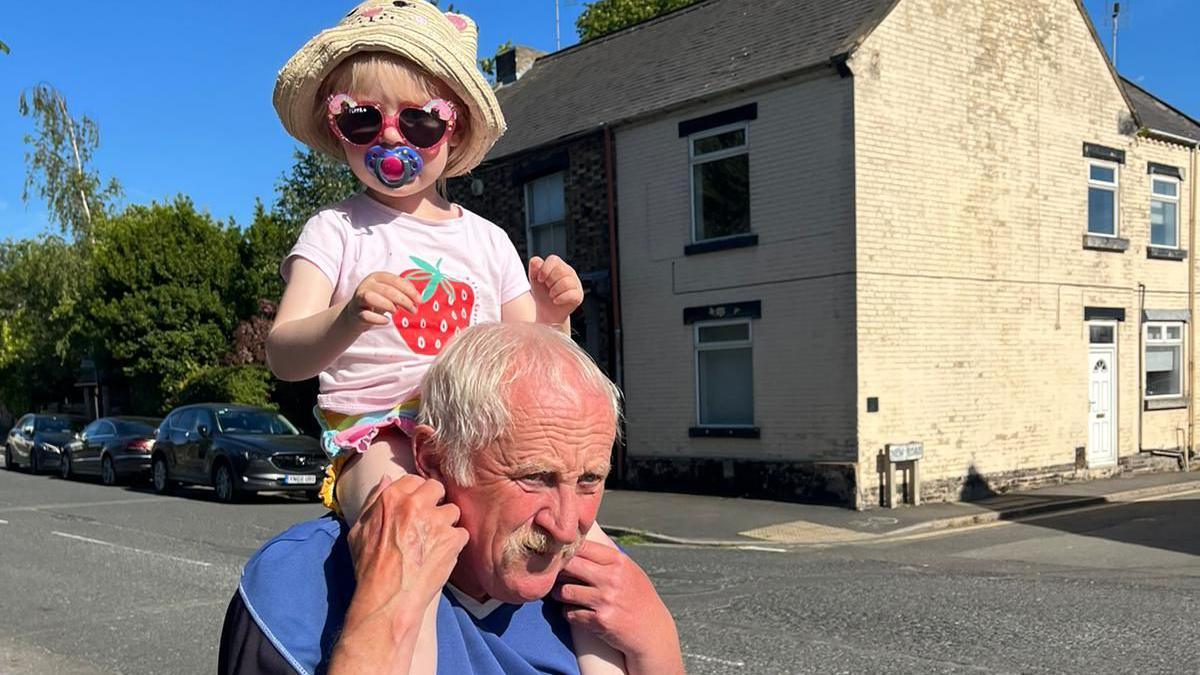 Philip Bramall, on a sunny residential street, with a small girl, wearing sunglasses and a straw hat, on his shoulders