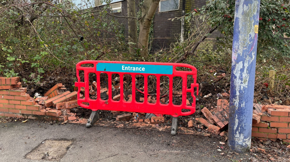 Pictured is a broken and collapsed red brick wall exposing trees and bushes. There is a red plastic barrier blocking off the hole in the wall.