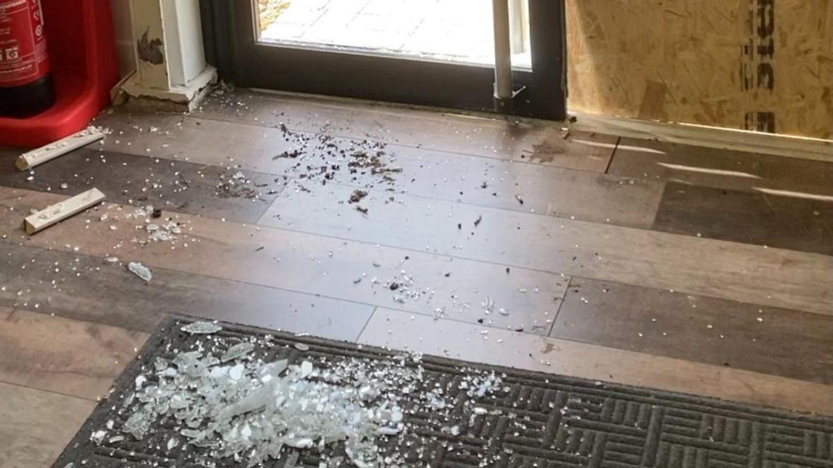 Glass on the floor, after a break-in showing glass on a mat, broken plastic, on a wood-effect floor. A door is in the distance.