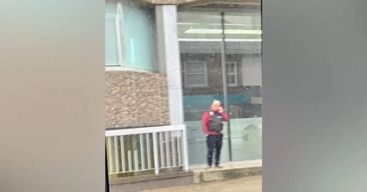 Blurry picture of a man wearing a tactical vest outside an office building. He is holding a mobile phone to his ear.